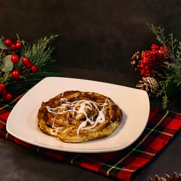 A cinnamon roll with white icing on a white plate, with festive holiday decorations.