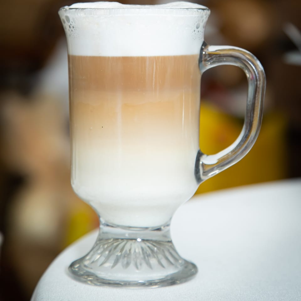 A tall glass mug of layered latte with frothy white foam on top.