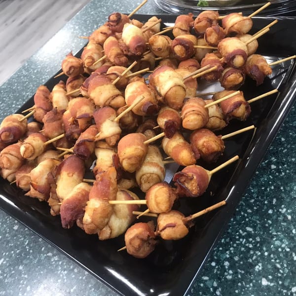 A black tray filled with numerous bacon-wrapped Brazilian appetizers on wooden skewers.
