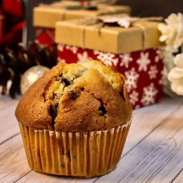A blueberry muffin sits on a light wooden surface, with festive gift boxes in the background.