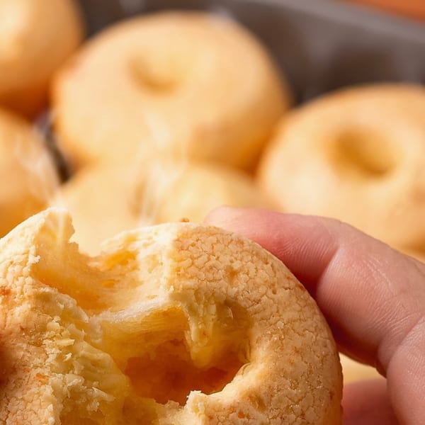 A hand holds a Brazilian cheese bread (pão de queijo) with a bite taken out, showing its fluffy interior.