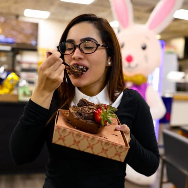 Woman in glasses enjoying a large chocolate dessert with strawberries, an Easter bunny in the background.