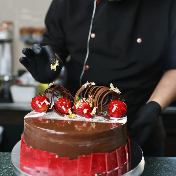 A chef in black gloves decorates a chocolate cake with strawberries and gold flakes.