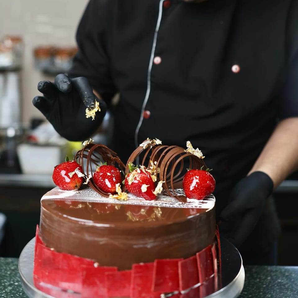 A chef in black gloves decorates a chocolate cake with strawberries and gold flakes.
