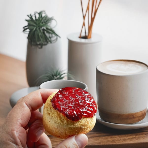 A hand holds a Brazilian pão de queijo topped with red guava paste, next to a latte.